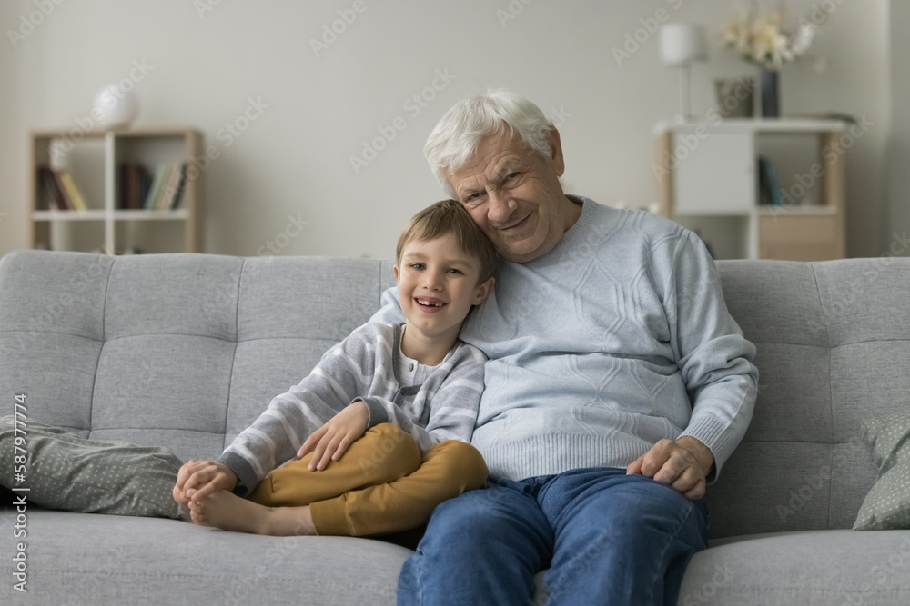Happy grandfather hugging cheerful cute preschool boy on home sofa, looking at camera, smiling, posing, enjoying visit of grandson kid, family leisure time. Grandpa and grandkid portrait