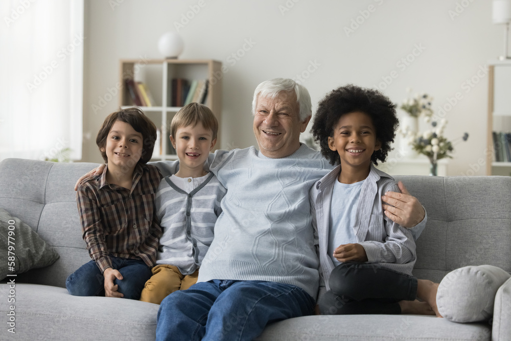 Happy diverse preschool kids and elder grandfather looking at camera, hugging, smiling, laughing, enjoying family diversity, friendship, leisure together, sitting on sofa. Front home portrait