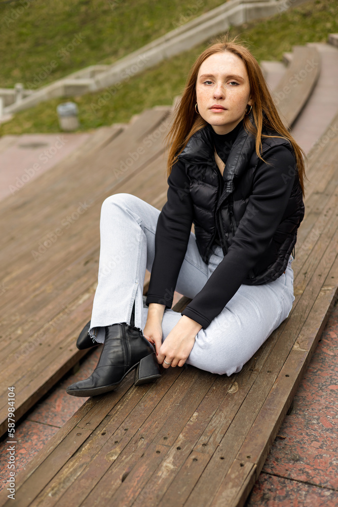 Young woman in black jacket and jeans sit on bench