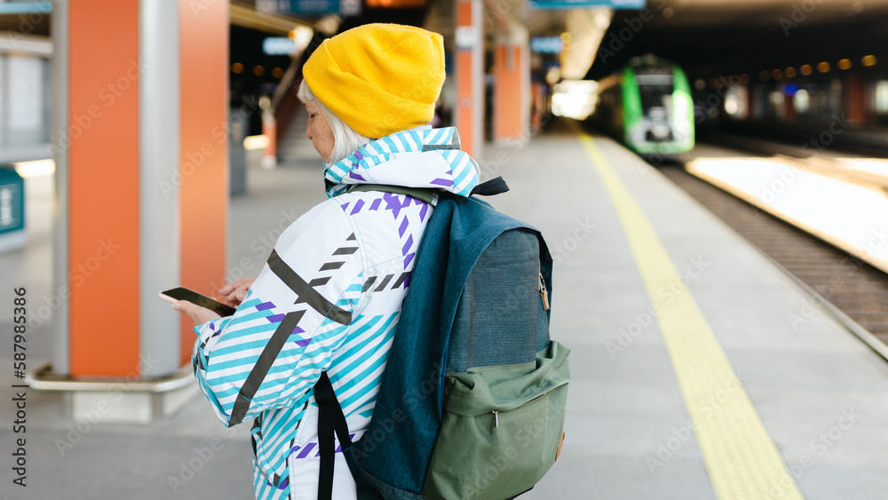 Old happy woman with backpack in yellow hat waiting train on station platform and using smart phone on urban background. Railroad transport concept Old happy woman with backpack in yellow hat waiting train on station platform and using smart phone on urban background. Railroad transport concept