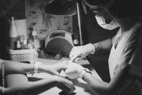 Manicurist female doctor in mask on face, making manicure procedure for hands with special equipment in the cabinet with mixed light, black&white image