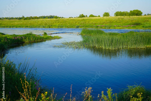 Sunny summer landscape with river. Green hills, fields and meadows. Quiet morning. Calm. Warm sunlight. Cloudless clear blue sky.