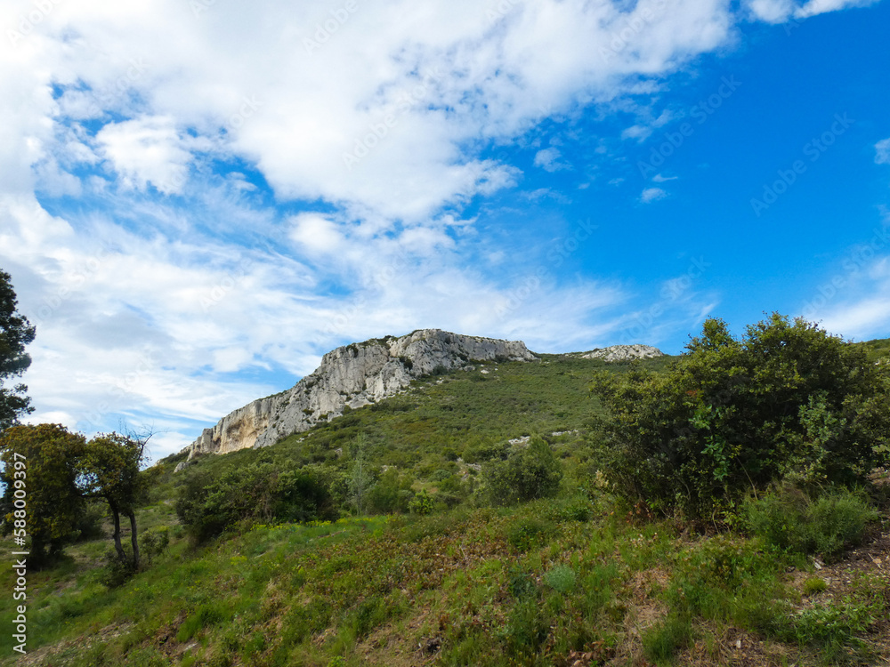 Natural landscape of the Alpilles in Provence in France where a rocky outcrop rises from a hill covered in scrubland under a beautiful blue sky adorned with pretty light fluffy white clouds