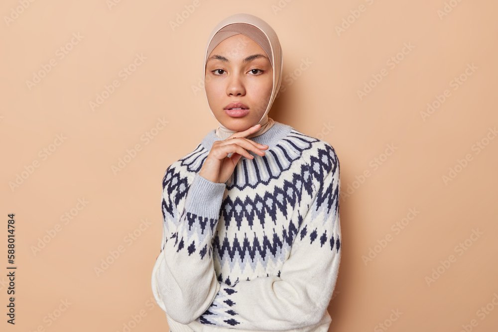 Studio shot of serious Muslim woman touches face gently looks directly at camera wears traditional hijab and sweater has calm expression isolated over brown background. People and religion concept Studio shot of serious Muslim woman touches face gently looks directly at camera wears traditional hijab and sweater has calm expression isolated over brown background. People and religion concept