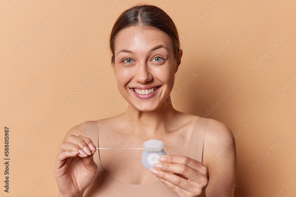 Teeth flossing concept. Dark haired smiling woman holds dental floss cares for perfect white teeth has healthy smooth skin dressed in t shirt stands bare shoulders isolated over brown background Teeth flossing concept. Dark haired smiling woman holds dental floss cares for perfect white teeth has healthy smooth skin dressed in t shirt stands bare shoulders isolated over brown background