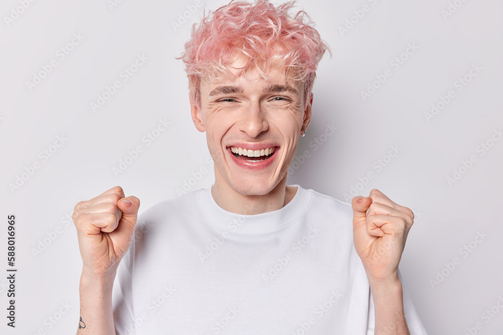 Enthusiastic happy hipster guy with trendy hairstyle clenches fists celebrates success smiles toothily dressed in casual basic t shirt isolated over white background glad to achieve great results Enthusiastic happy hipster guy with trendy hairstyle clenches fists celebrates success smiles toothily dressed in casual basic t shirt isolated over white background glad to achieve great results