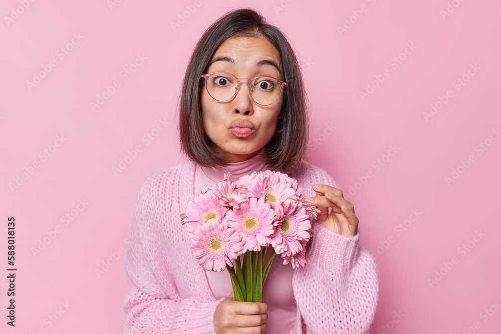 Portrait of surprised Asian woman with dark hair keeps lips folded holds bouquet of gerbera flowers wears knitted sweater and transparent eyeglasses isolated over pink background. Monochrome shot Portrait of surprised Asian woman with dark hair keeps lips folded holds bouquet of gerbera flowers wears knitted sweater and transparent eyeglasses isolated over pink background. Monochrome shot