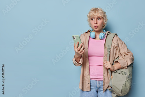 Horizontal shot of stunned femalee student with rucksack holds mobile phone reacts to shocking news dressed in shirt and jeans isolated over blue background copy space for your promotional content photo