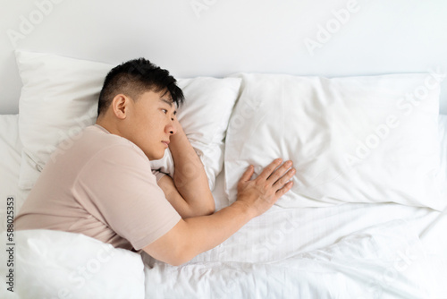 Lonely asian man lying in bed, touching empty pillow © Prostock-studio