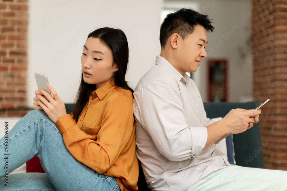 Marital crisis, indifference. Korean spouses using cellphones, sitting back-to-back on sofa, indifferent to each other