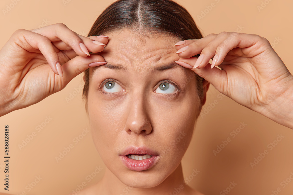 Fototapeta premium Close up shot of brunette young woman touches forehead worried about skin wrinkles needs anti aging treatment focused above isolated over brown background. Health beauty and dermatology concept