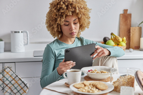 Serious curly woman uses digital tablet watches film while having breakfast or surfs internet sits at table with cereals and cup of coffee against kitchen interior dressed in domestic clothing