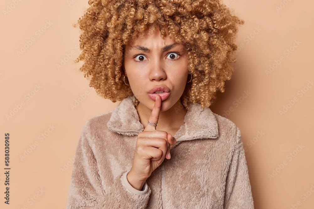 Serious strict woman with curly hair makes silence gesture demands to keep information in secret has scared surprised expression wears jacket isolated over brown background. Dont drop any word Serious strict woman with curly hair makes silence gesture demands to keep information in secret has scared surprised expression wears jacket isolated over brown background. Dont drop any word