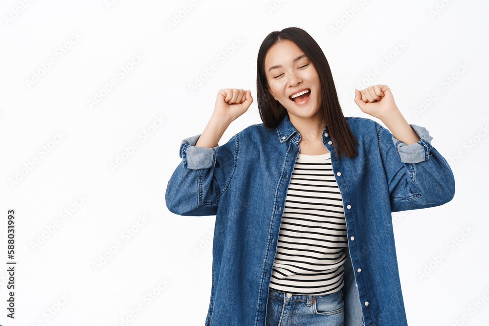 Enthusiastic asian girl dancing and triumphing, raising fists up and chanting yes, celebrating victory, standing over white background Enthusiastic asian girl dancing and triumphing, raising fists up and chanting yes, celebrating victory, standing over white background