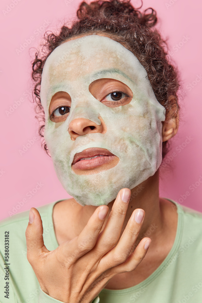 Fototapeta premium Headshot of serious woman applies moisturising sheet mask does skin care procedures touches face looks attentively at camera isolated over pink background background. Beauty procedures concept
