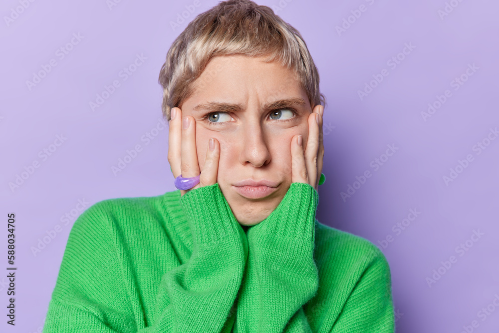 Indoor shot of displeased short haired woman keeps hands on cheeks has sad gloomy offended expression wears loose green jumper isolated over purple background. Negative facial expressions concept Indoor shot of displeased short haired woman keeps hands on cheeks has sad gloomy offended expression wears loose green jumper isolated over purple background. Negative facial expressions concept