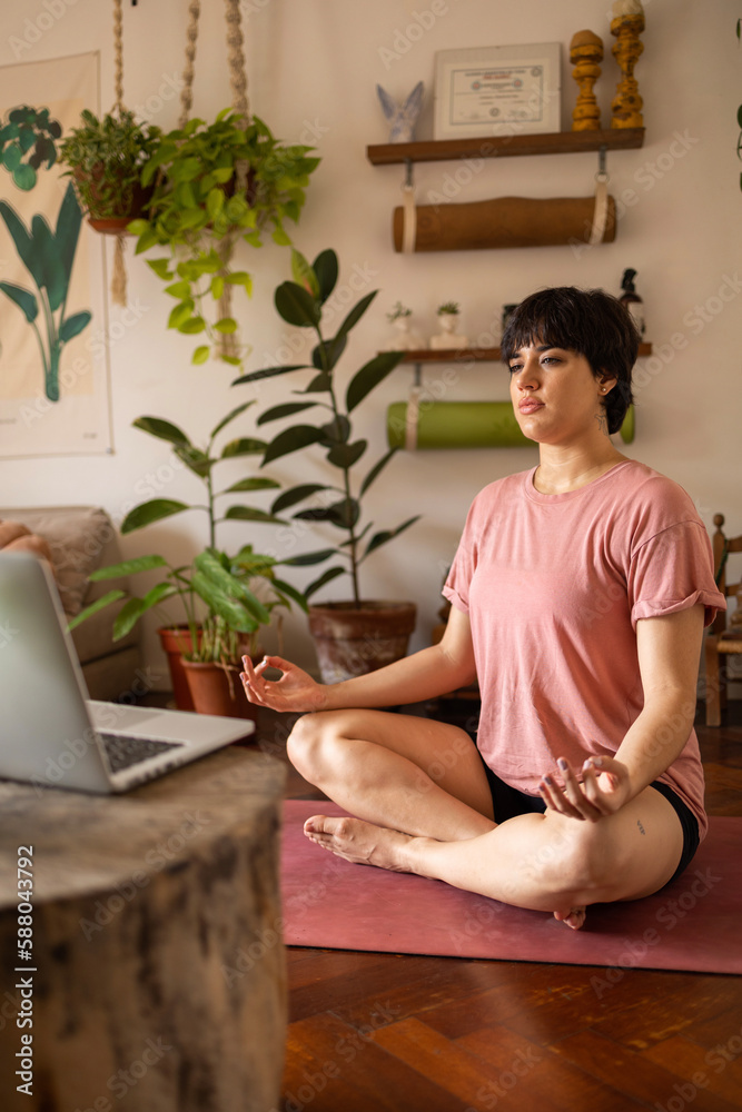 Vertical of young latin woman with a short hair meditating while looking at the laptop. She is wearing pink shirt and sitting in the room full of green plants.