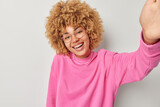 Selfie shot of positive cheerful woman poses for photograph keeps arm outstretched smiles broadly dressed in casual pink pullover transparent eyeglasses isolated on white background stands in studio.