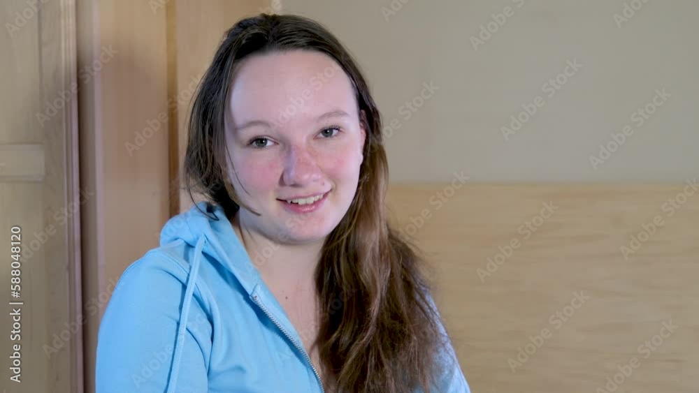 teenage girl with unwashed hair in a room in a blue blouse looks into the frame shakes her head in agreement smiling round face of a teenager teenage girl with unwashed hair in a room in a blue blouse looks into the frame shakes her head in agreement smiling round face of a teenager