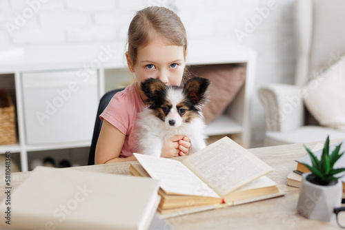 cute little preschool girl reading book at home with her puppy papillon dog