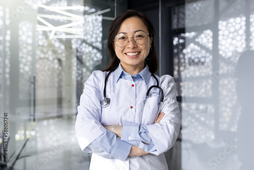 Portrait of a young beautiful and successful Asian woman inside a clinic office, a doctor smiling and looking at the camera with crossed arms and a white medical coat with a stethoscope. © Liubomir