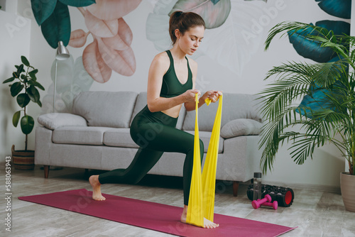 Full body young strong sporty athletic fitness trainer instructor woman wears green tracksuit using elastic rubber band training doing exercises at home gym indoor. Workout sport motivation concept.