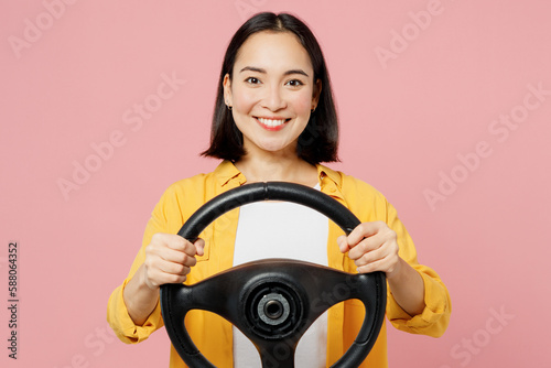Young happy fun woman of Asian ethnicity wear yellow shirt white t-shirt hold steering wheel driving car look camera isolated on plain pastel light pink background studio portrait. Lifestyle concept.
