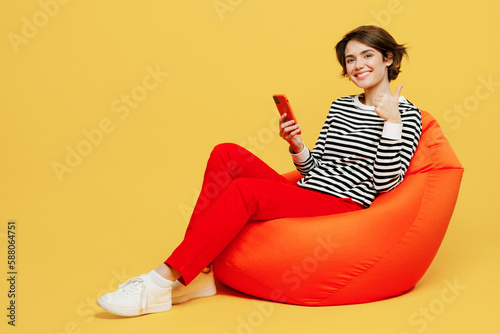 Full body young woman wear casual black and white shirt sit in bag chair hold in hand use mobile cell phone show thumb up isolated on plain yellow color background studio portrait. Lifestyle concept.