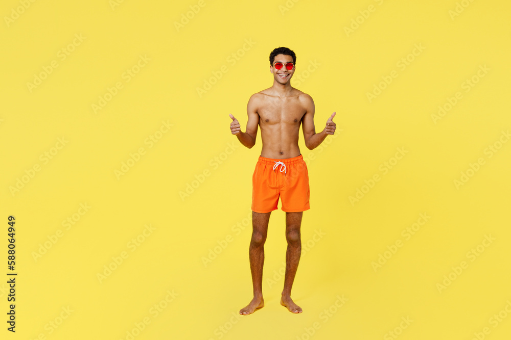 Full body satisfied smiling young sexy man wear orange shorts swimsuit relax near hotel pool showing thumb up like gesture isolated on plain yellow background Summer vacation sea rest sun tan concept