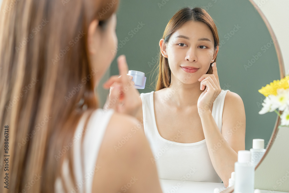 Fresh skin, beautiful smile of asian young woman, girl looking at mirror, hand applying moisturizer lotion on her face, putting cream treatment before makeup cosmetic routine at home. Facial Beauty. Fresh skin, beautiful smile of asian young woman, girl looking at mirror, hand applying moisturizer lotion on her face, putting cream treatment before makeup cosmetic routine at home. Facial Beauty.