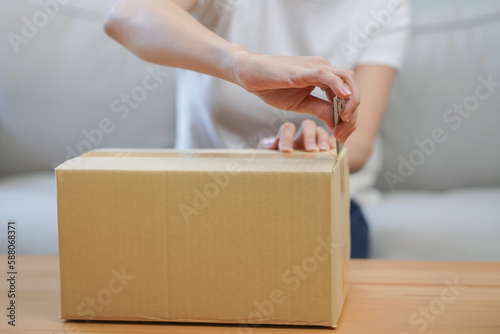 Happy asian young woman, girl customer sitting on sofa at home, hand in opening and unpacking cardboard box carton parcel after buying ordering present, shopping online, delivery service concept.