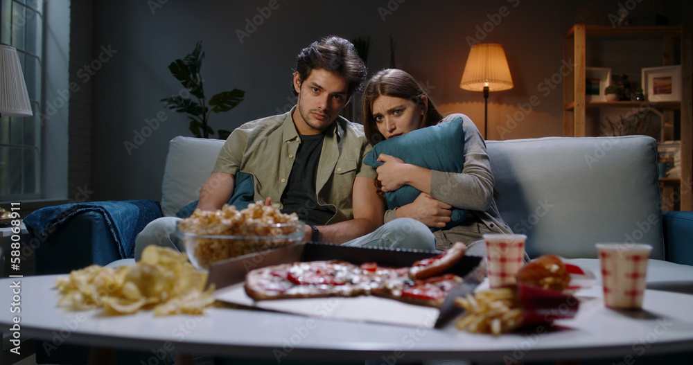 Young caucasian couple spending time in front of tv together, being scared by a horror movie and eating unhealthy food 