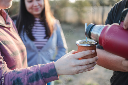  Close up of smiling friends drinking yerba mate using a thermos with hot water in the countryside at sunset. photo