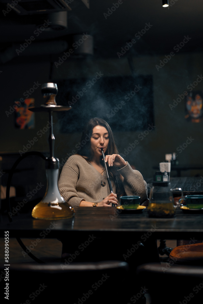 Naklejka premium Beautiful young woman inhaling hookah girl smoking hookah sitting in lounge bar concept of relaxation and smoking