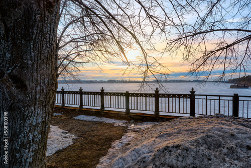 View of the setting sun through the fence and tree branches near the banks of the Volga River. Spring sunset near the river. The last spring snow on the river bank. © Lexis_Jan