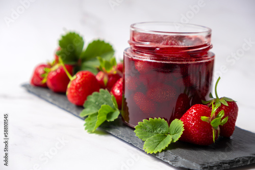 Homemade strawberry preserves or jam in glass jar on slate dish with fresh strawberry fruit and green leaves on marble. Recipe of delicious homemade berry jam of strawberry full of vitamins.