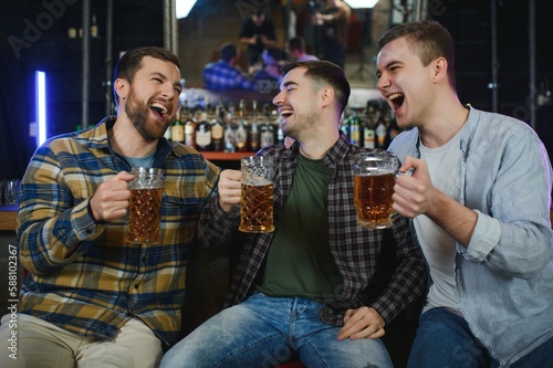 Three young men in casual clothes are smiling and clanging glasses of beer together while sitting at bar counter in pub