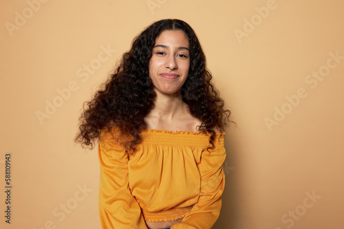 Portrait of charming young lady of 20s with dark skin and black long wavy hair in yellow summer top looking at camera with shy facial expression and timid smile on brown studio background