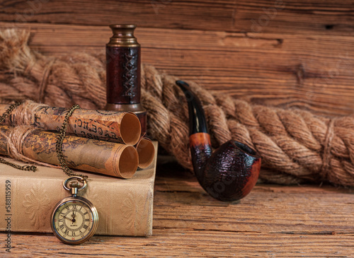Stare, eleganckie przedmioty, zwoje map i zegarek kieszonkowy leżące na książce oraz luneta, fajka do tytoniu i gruba lina na drewnianym tle. 