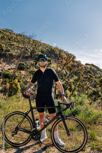 Male cyclist with bicycle in wild terrain looking at camera. Athlete in black sportswear with road bike relaxing.