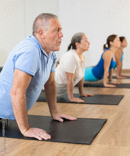 Focused aged man doing yoga in group of adults in modern studio, standing in upward facing dog pose. Healthy sports lifestyle concept