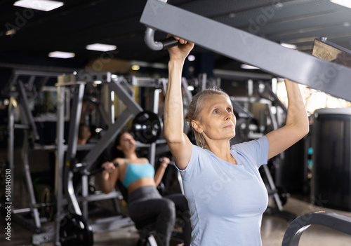 Portrait of motivated senior woman performing set of exercises for back, doing lat pull-down on lever gym machine. Concept of weight training for older adults..