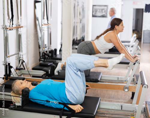Focused elderly woman lying while doing Pilates tone exercise for back on reformer bed equipment in gym