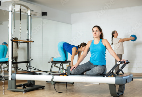 Positive concentrated young girl practicing pilates stretching exercises on reformer at gym..