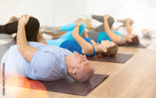 Concentrated elderly man doing stretching exercises during group training in gym, lying on mat and pulling knee toward shoulder to relax glutes and release tension from sciatic nerve..