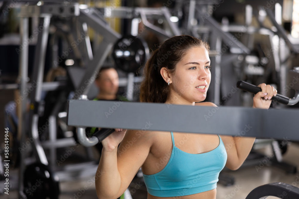 Fototapeta premium Sporty young woman using shoulder press machine in gym