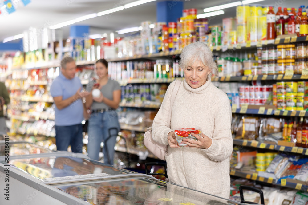 Slender elderly woman in casual knitted dress shopping for groceries in food store, looking at products in glass refrigerator with interest