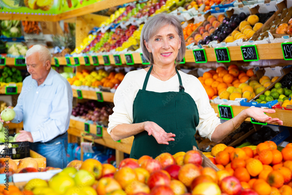Mature woman greengrocer worker standing in salesroom and looking in camera. Old man customer shopping in background.