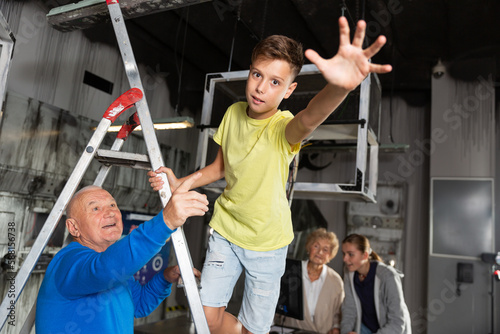 Young boy standing on stepladder and reaching his hand in escape room. His grandfather standing next to him and pointing.