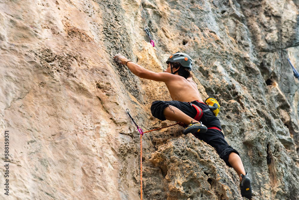 Obraz premium Young Asian man climber climbing on rocky coastline at tropical island in sunny day. Handsome guy enjoy outdoor active lifestyle and extreme sport training mountain climbing on summer holiday vacation